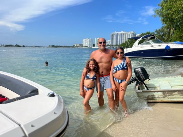 Smiling father with two daughters in matching blue-striped swimsuits standing in shallow turquoise water beside anchored boats and waterfront condos under a sunny blue sky