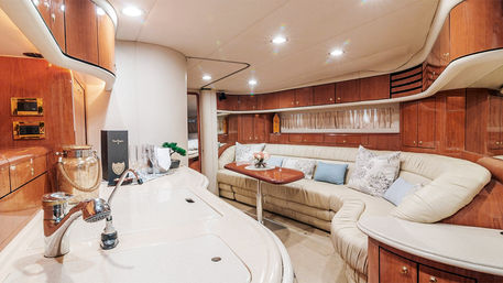 Luxury yacht interior: cozy cream leather L-shaped salon with glossy wood cabinets, compact galley sink, small wooden table with flowers and champagne flutes, and warm recessed lighting