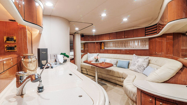 Luxury yacht interior: cozy cream leather L-shaped salon with glossy wood cabinets, compact galley sink, small wooden table with flowers and champagne flutes, and warm recessed lighting