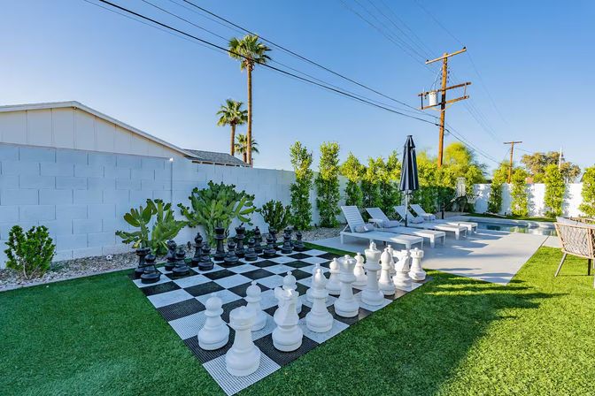 Resort-style sunny backyard with giant outdoor chess set on black-and-white checkerboard turf beside a pool, white lounge chairs, palm trees and a concrete patio