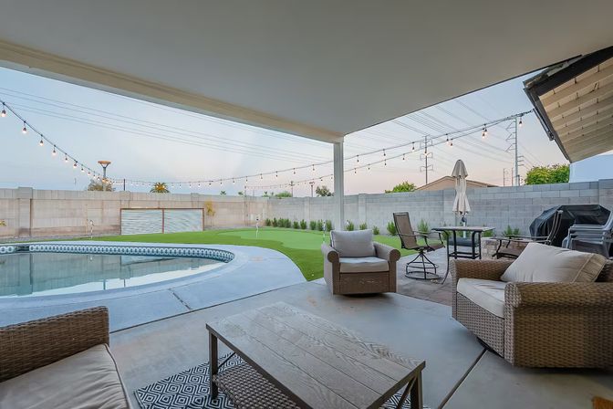 Cozy covered backyard patio with wicker seating and coffee table overlooking a curved swimming pool, small putting green, patio dining set, grill and string lights at dusk.