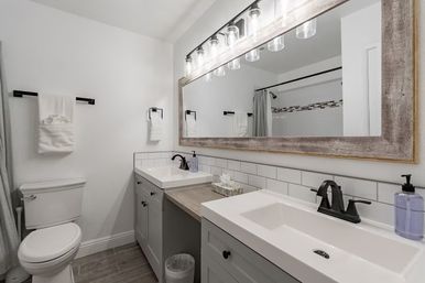 Bright modern home bathroom with double white sinks, gray shaker vanities, large wood‑framed mirror, subway tile backsplash, black matte faucets, toilet and shower with curtain