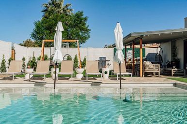 Sunny residential backyard pool with turquoise water, beige lounge chairs and closed white umbrellas, pergola-covered outdoor seating, palm tree and white privacy wall under a clear blue sky