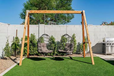 Sunny suburban backyard with a wooden A-frame holding two hanging egg chairs over green turf, white cinderblock wall, leafy shrubs and a stainless-steel grill under a clear blue sky.