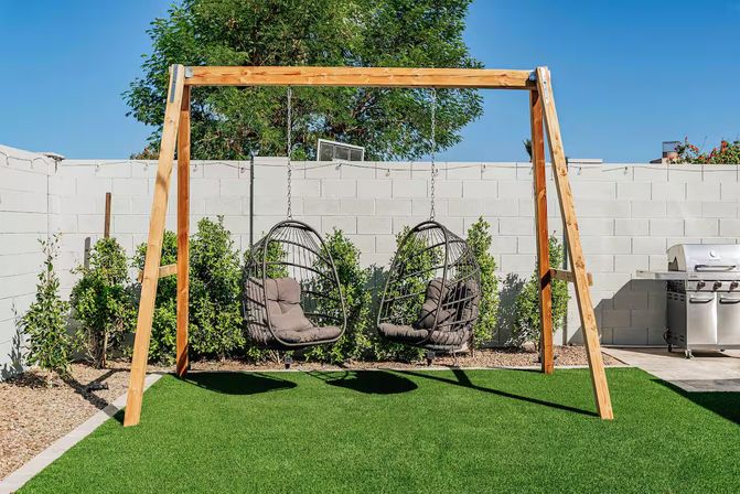 Sunny suburban backyard with a wooden A-frame holding two hanging egg chairs over green turf, white cinderblock wall, leafy shrubs and a stainless-steel grill under a clear blue sky.