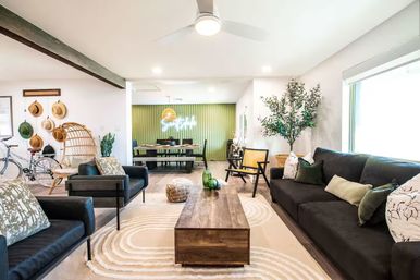 Bright open-concept living room with dark sofas, wooden coffee table on a circular-pattern rug, rattan accent chair, bicycle and hat wall, indoor tree, and a green-accent dining area in the back.