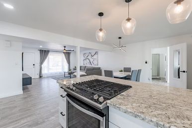 Bright, airy open-concept kitchen and living area with granite island and stainless steel gas range, glass pendant lights, gray tile floors, dining table and large curtained window.