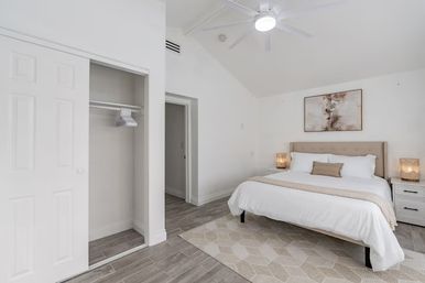Bright airy modern master bedroom with king bed and tufted beige headboard, white bedding, nightstands with woven lamps, abstract wall art, ceiling fan, open closet, wood-look tile floor and neutral geometric rug