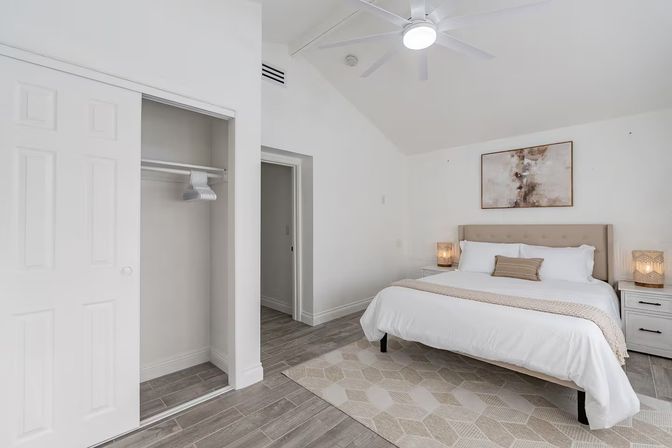 Bright airy modern master bedroom with king bed and tufted beige headboard, white bedding, nightstands with woven lamps, abstract wall art, ceiling fan, open closet, wood-look tile floor and neutral geometric rug