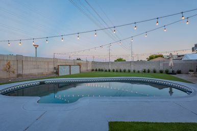 Suburban backyard at dusk featuring a kidney-shaped pool reflecting hanging string lights, concrete patio, small putting green, and cinderblock privacy wall with power lines overhead.