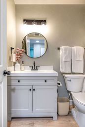 Cozy modern small bathroom with gray vanity and white countertop, round mirror under three-bulb light, black faucet, folded white towels, pink floral vase and toilet.