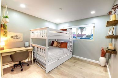 Bright modern kids' bedroom with white bunk bed and trundle, light wood desk and swivel chair, pale wood floors, decorative pillows and 'Wild West' wall art, hanging plants, floating shelves and a window with a palm tree view.