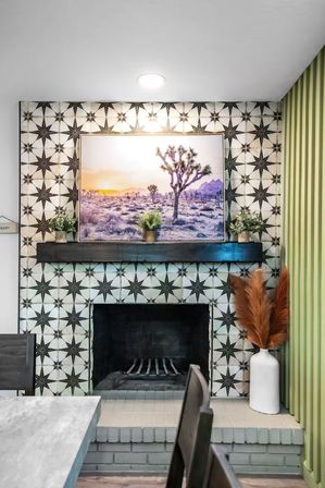 Cozy modern fireplace framed by black star-pattern tiles, dark wood mantel with a desert sunset print and potted plants, and a white vase of rust-colored pampas grass.