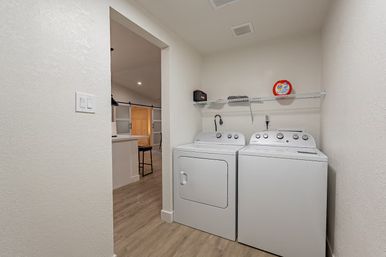 Compact bright laundry nook with white top‑load washer and matching dryer side-by-side beneath a wire shelf holding laundry supplies, wood‑look flooring, and an open doorway to the adjacent kitchen.
