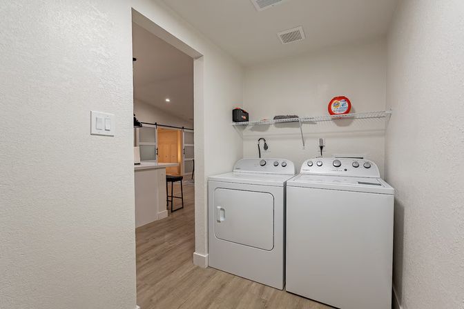 Compact bright laundry nook with white top‑load washer and matching dryer side-by-side beneath a wire shelf holding laundry supplies, wood‑look flooring, and an open doorway to the adjacent kitchen.