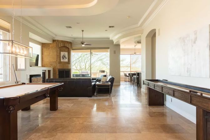 Open-plan luxury game room and living area with pool table, shuffleboard, leather sectional, fireplace and large windows framing a sunny desert landscape and natural stone tile floors.