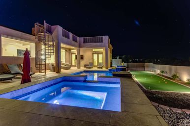 Nighttime view of a modern luxury backyard with a glowing blue pool and spa, outdoor lounge seating, spiral staircase to an upper deck, and a lit artificial putting green under a starry sky.