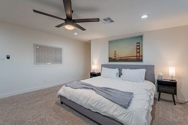 Cozy modern master bedroom with king bed, gray upholstered headboard, white bedding and blue throw, ceiling fan, bedside tables with lamps, plush carpet, and framed Golden Gate Bridge wall art.