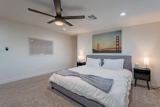 Cozy modern master bedroom with king bed, gray upholstered headboard, white bedding and blue throw, ceiling fan, bedside tables with lamps, plush carpet, and framed Golden Gate Bridge wall art.
