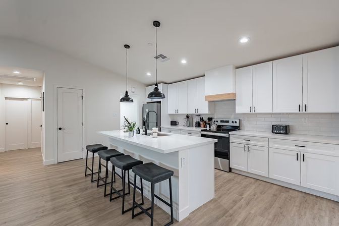 Bright modern white kitchen with a long island and four black bar stools, stainless-steel appliances, black pendant lights, subway tile backsplash, and light wood floors.