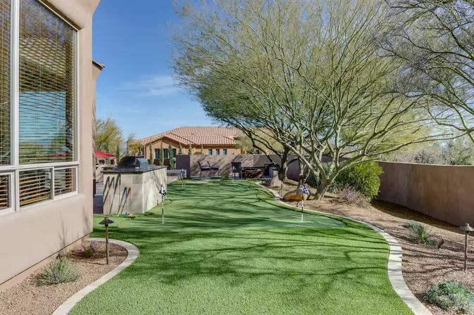 Sunlit Southwestern desert backyard with artificial putting green, built-in outdoor grill island, practice flags, mature desert trees and stucco walls under a blue sky.