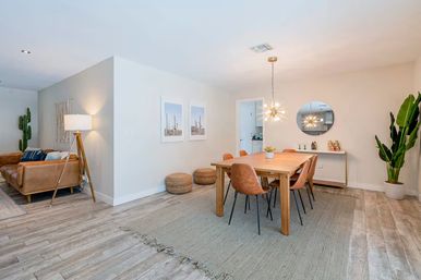 Bright modern open-plan dining room and living area with a wooden table and six tan leather chairs on a large woven rug, starburst pendant light and round mirror, potted plants, cactus wall art and a leather sofa with floor lamp on light wood flooring.