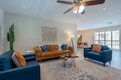 Sunlit boho-modern living room with tan leather sofa, two blue armchairs, patterned area rug, live cactus, wood-slab coffee table, macramé wall hanging, ceiling fan, and dining area by shuttered windows.