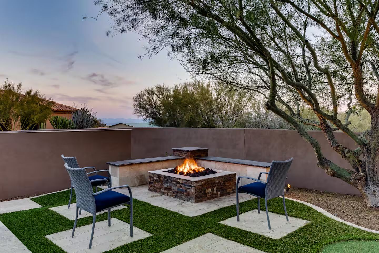 Cozy desert backyard patio at dusk with a lit stone fire pit surrounded by four woven chairs, stucco privacy wall, native trees, and patterned turf