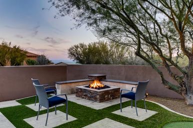Cozy desert backyard patio at dusk with a lit stone fire pit surrounded by four woven chairs, stucco privacy wall, native trees, and patterned turf
