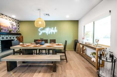 Bright modern boho dining room in Scottsdale with a long wooden table and bench set for six, rattan pendant light, green vertical slat accent wall with neon script sign, patterned tile fireplace, large window, wooden console and bar cart.