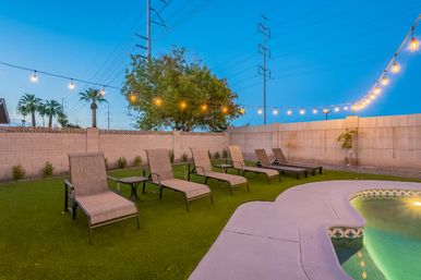 Twilight suburban backyard pool scene with glowing string lights, a row of chaise lounges on green turf, palm trees and a masonry wall under a clear blue sky — inviting evening oasis.