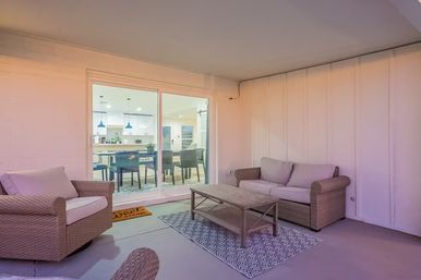 Cozy covered patio with wicker sofa and armchairs, coffee table on a patterned rug, and sliding glass door opening to a bright modern kitchen and dining area.