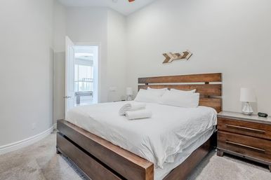 Bright modern master bedroom interior with king-size wooden platform bed, crisp white linens and folded towels, matching wood nightstands and lamps, neutral walls, soft gray carpet and an open doorway to an en-suite bathroom.