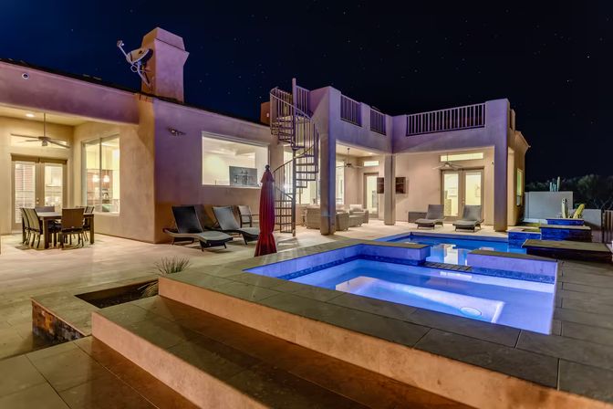 Nighttime view of a modern luxury backyard: glowing blue pool and spa, lounge chairs and outdoor dining, spiral staircase to a rooftop deck under a starry sky.