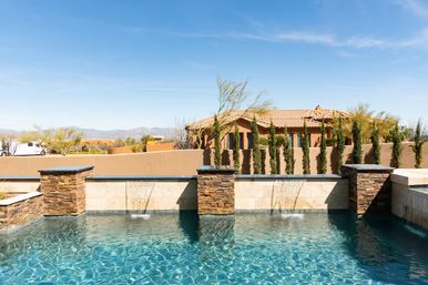 Sunny backyard pool with turquoise water and three stone waterfall features, framed by cypress trees and a stucco Southwestern house with clay tile roof and desert mountains under a clear blue sky.