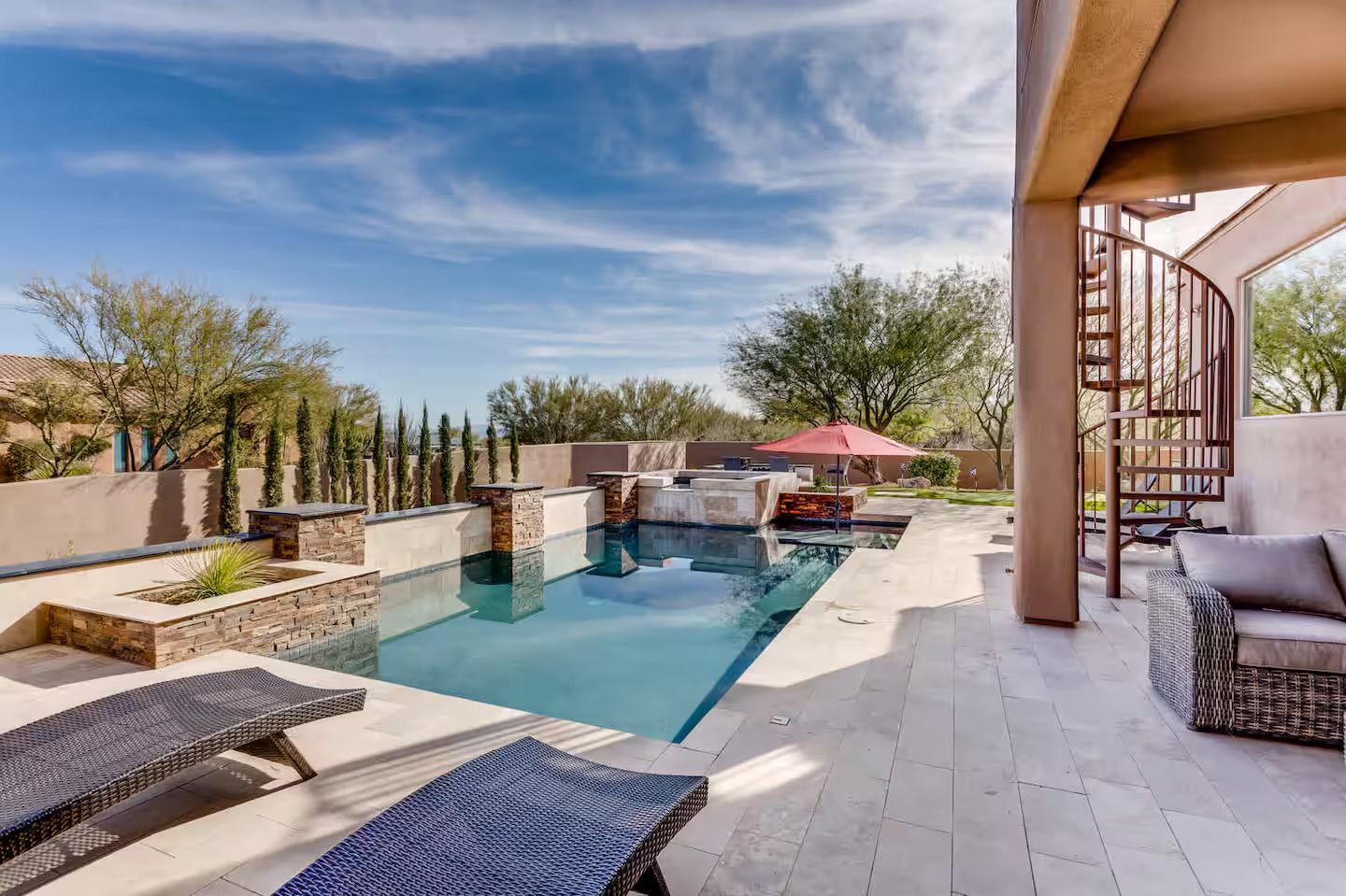 Sunny southwestern backyard oasis with a modern rectangular pool and raised stone spa, water features, wicker lounge chairs, red patio umbrella, spiral staircase and desert landscaping.