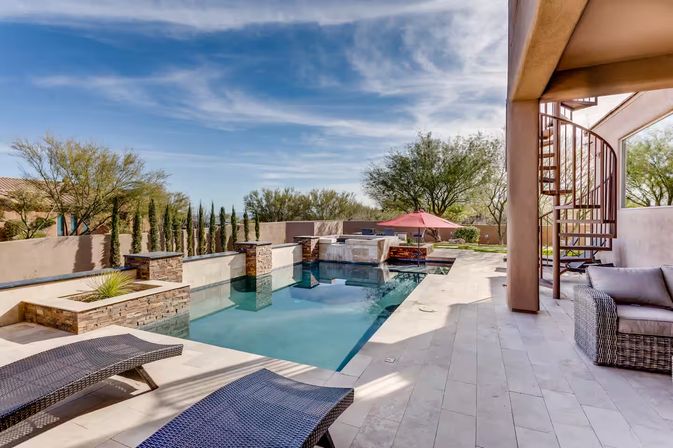 Sunny southwestern backyard oasis with a modern rectangular pool and raised stone spa, water features, wicker lounge chairs, red patio umbrella, spiral staircase and desert landscaping.