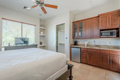 Sunlit studio bedroom with kitchenette — white bed linens, TV on a dresser, ceiling fan, wooden cabinets with microwave and mini-fridge, tiled floor.