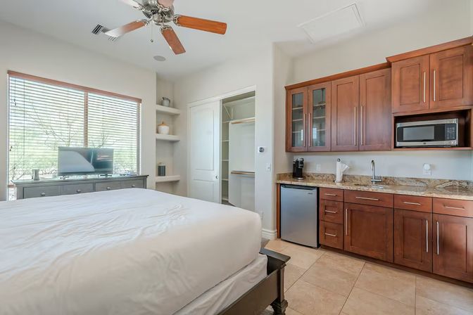 Sunlit studio bedroom with kitchenette — white bed linens, TV on a dresser, ceiling fan, wooden cabinets with microwave and mini-fridge, tiled floor.