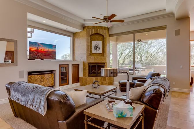 Cozy modern living room with dark leather sofas, tiled corner fireplace, coffee table, TV showing a desert sunset, and large sliding glass doors opening to a sunlit patio with trees.