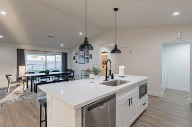 Bright modern open-plan kitchen and dining area with white quartz island, undermount sink and black gooseneck faucet, stainless dishwasher, two black pendant lights, long wooden dining table with bench seating by a large sliding glass door, light wood floors and neutral walls.