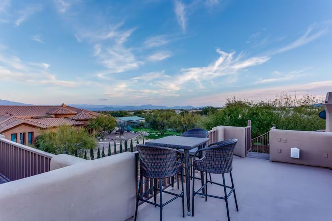 Rooftop patio with wicker bar table and four stools, stucco railing, desert landscaping and distant mountains under a pastel sunset sky.