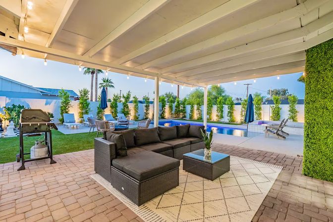Covered backyard patio with wicker sectional and rug under string lights, overlooking a blue swimming pool, lounge chairs, grill and palm trees.