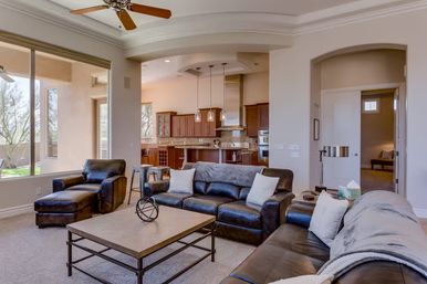 Open-concept living room and kitchen with dark leather sofas, chaise lounge and ottoman, metal-framed coffee table, large windows with natural light, ceiling fan, and kitchen island with pendant lights and wood cabinetry