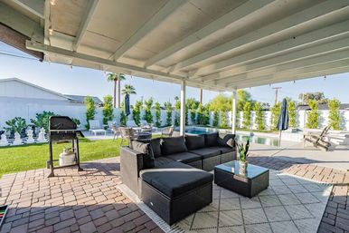 Sunny covered backyard patio with black wicker sectional and coffee table on a patterned rug, propane grill on paver patio, green lawn, pool with lounge chairs and palm trees in a suburban yard