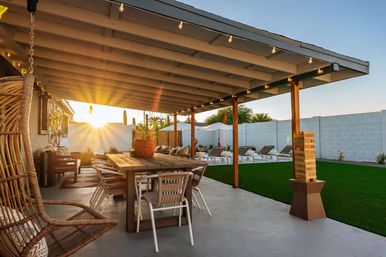 Sunset-covered backyard patio with wooden dining table, rattan hanging chair, string lights, green lawn and sun loungers under umbrellas