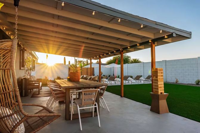 Sunset-covered backyard patio with wooden dining table, rattan hanging chair, string lights, green lawn and sun loungers under umbrellas