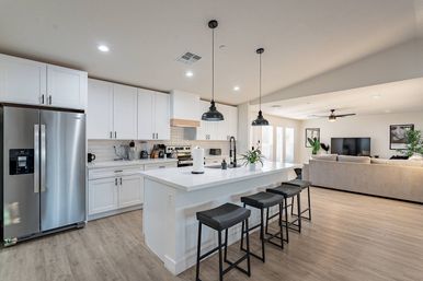 Sleek open-concept kitchen and living area with a large white island and quartz countertop, black pendant lights over four bar stools, stainless steel fridge and appliances, white shaker cabinets, subway tile backsplash, light wood floors, and a bright beige sofa in the adjacent living room.