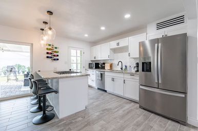 Sunlit modern open-plan white kitchen with large island and granite countertop, four black swivel bar stools, stainless steel French-door refrigerator and gas cooktop, globe pendant lights, wall wine rack and sliding patio door to outdoor seating.