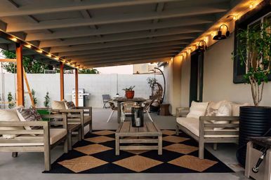 Covered backyard patio with wooden lounge seating, geometric rug, string lights, dining table, hanging rattan chair, potted plants and a grill